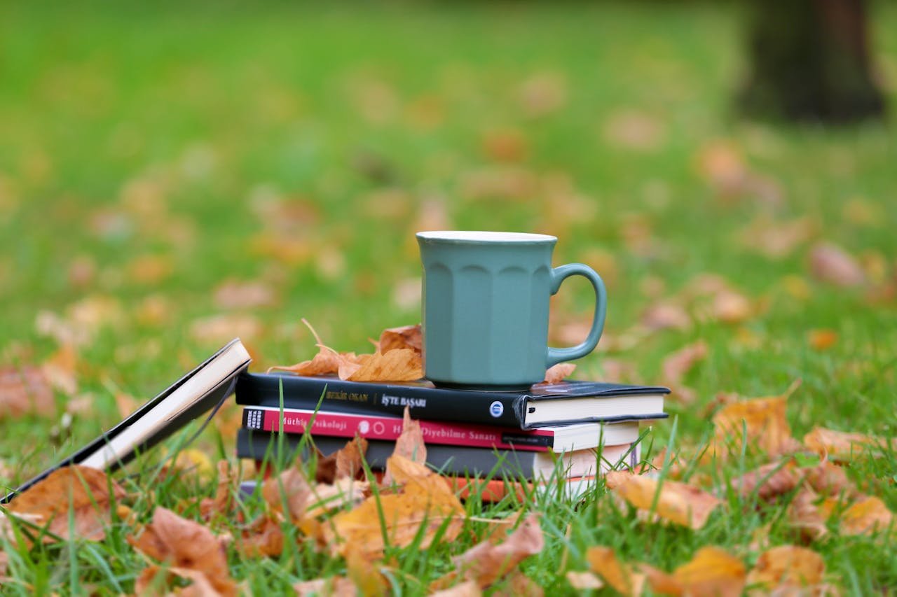 Home A serene autumn setup of books and a cup on grassy field with fallen leaves.