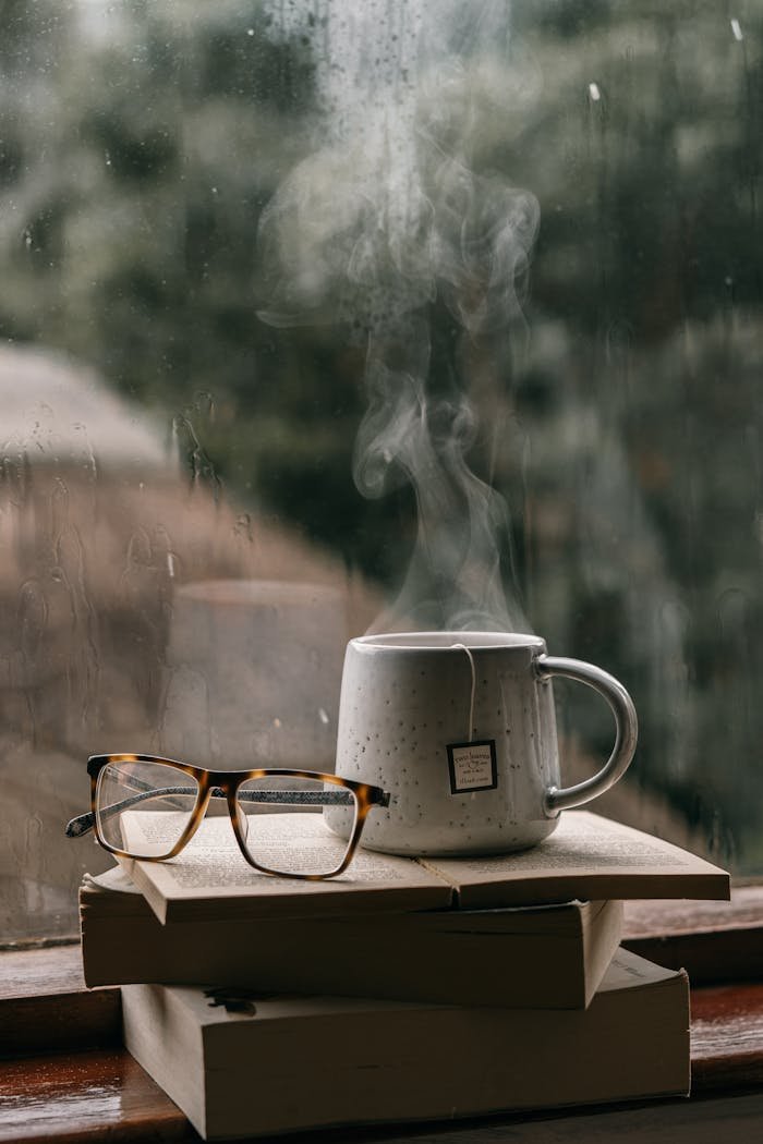 Home Comforting setup with books, glasses, and hot tea by a rain-drenched window, perfect for reading.